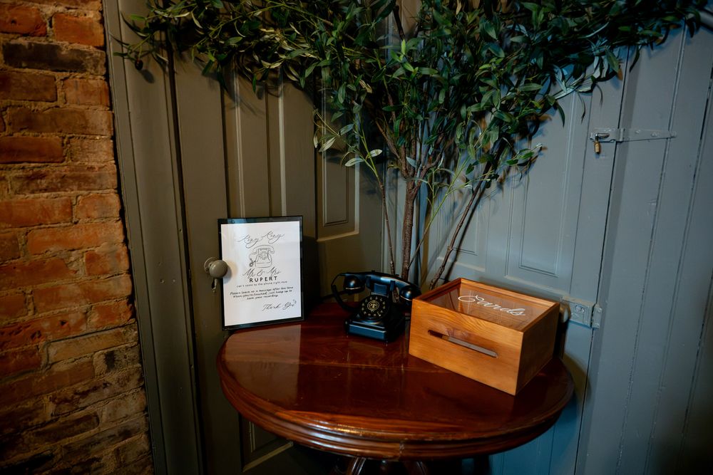 Vintage telephone and wooden box on round mahogany table beside brick wall and olive tree.