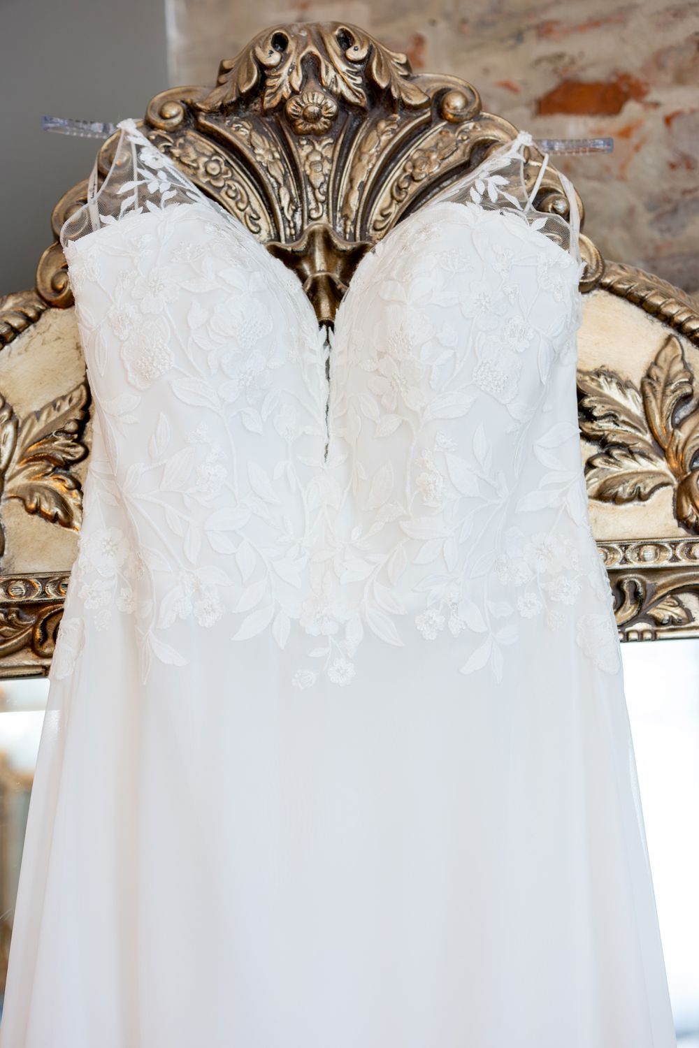 White lace wedding dress with sweetheart neckline hanging on ornate gold mirror frame.
