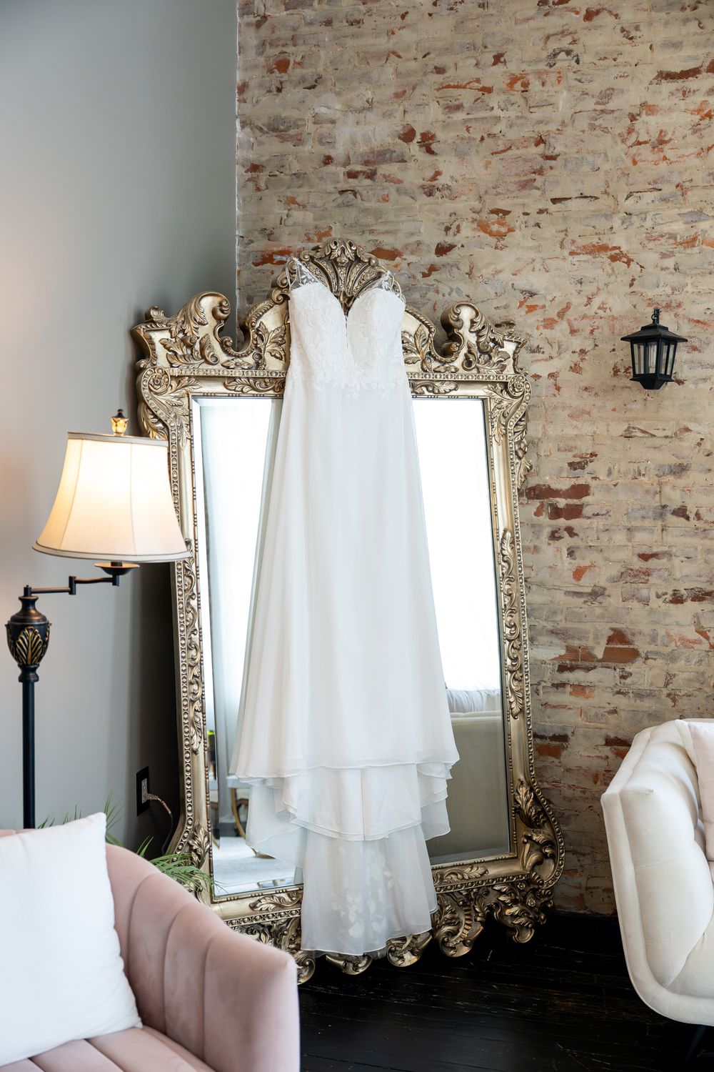 White wedding dress hanging on ornate gold mirror against exposed brick wall with lamp and seating nearby.