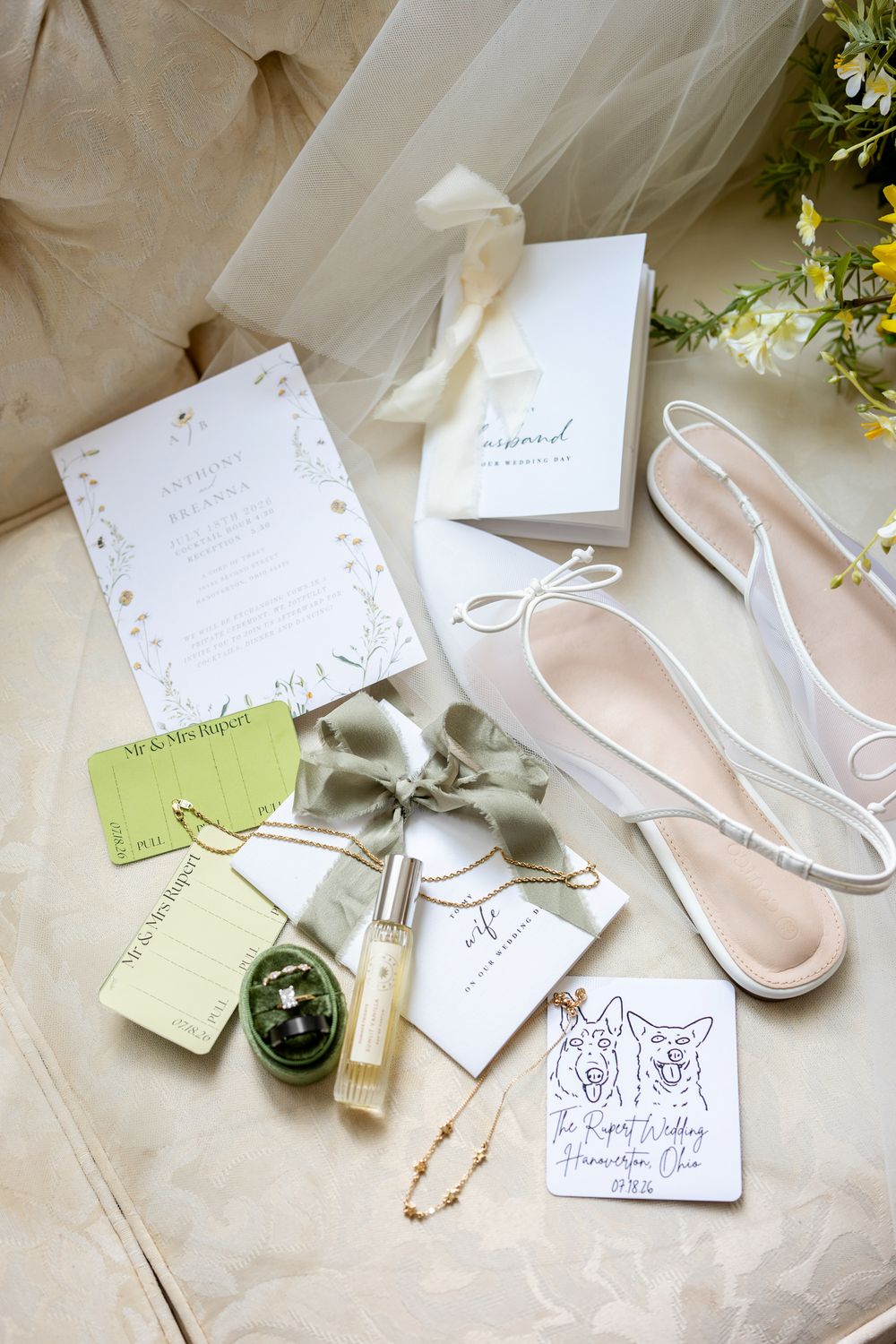 Elegant wedding flat lay featuring floral invitations, blush satin shoes, perfume, green jewelry box, and silk ribbon on beige surface.