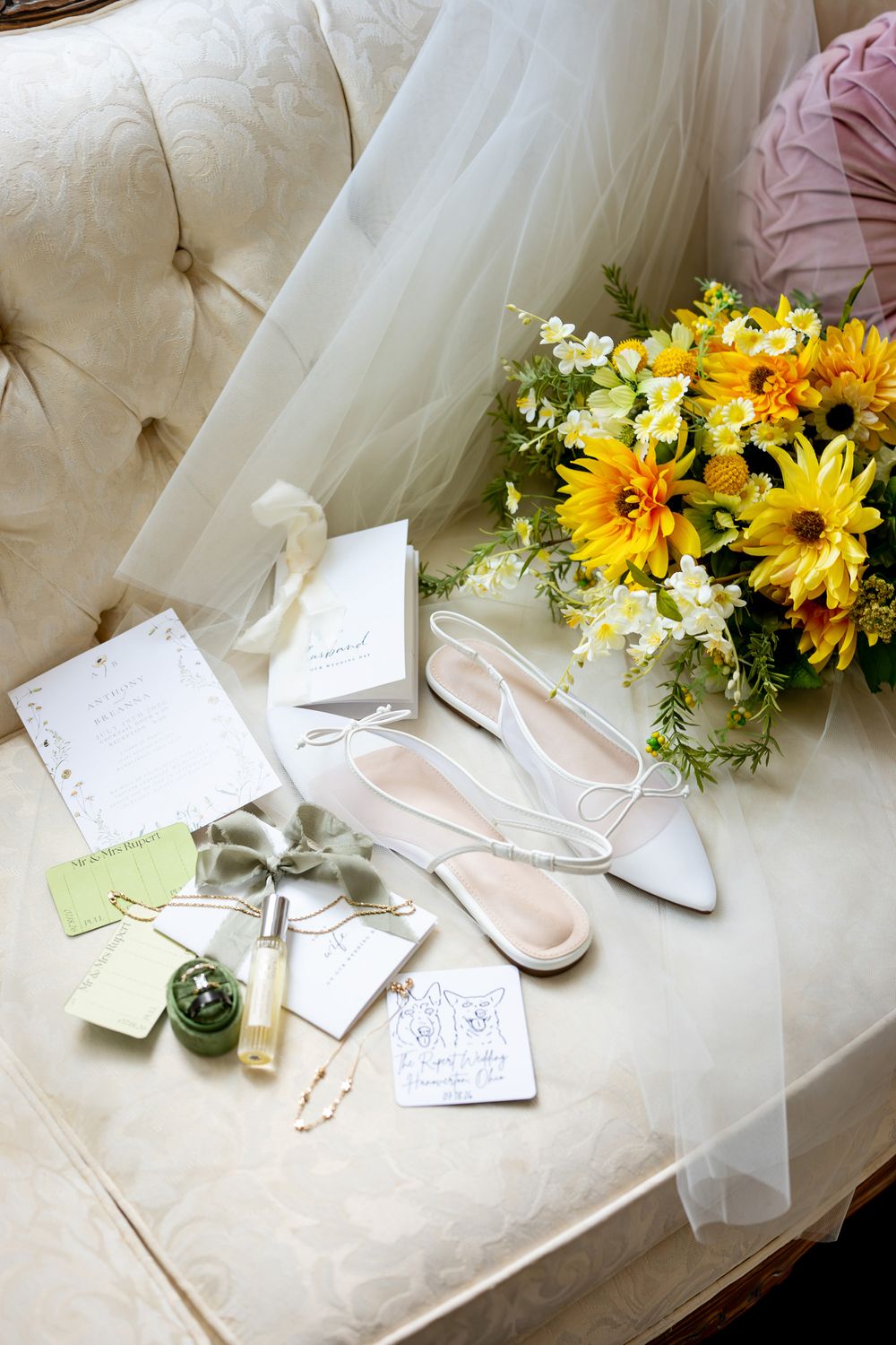 Bridal flat shoes, sunflower bouquet, perfume, and wedding stationery arranged on a cream tufted chair.