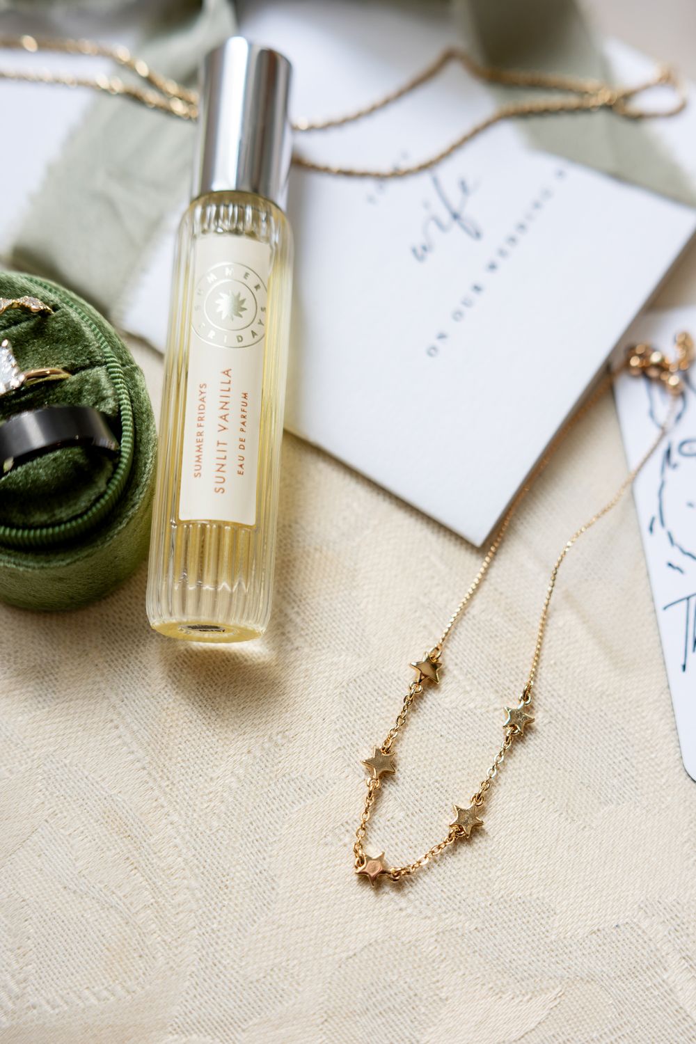 Flat lay of sunlit vanilla perfume, gold star necklace, ring box, and wedding card on linen.