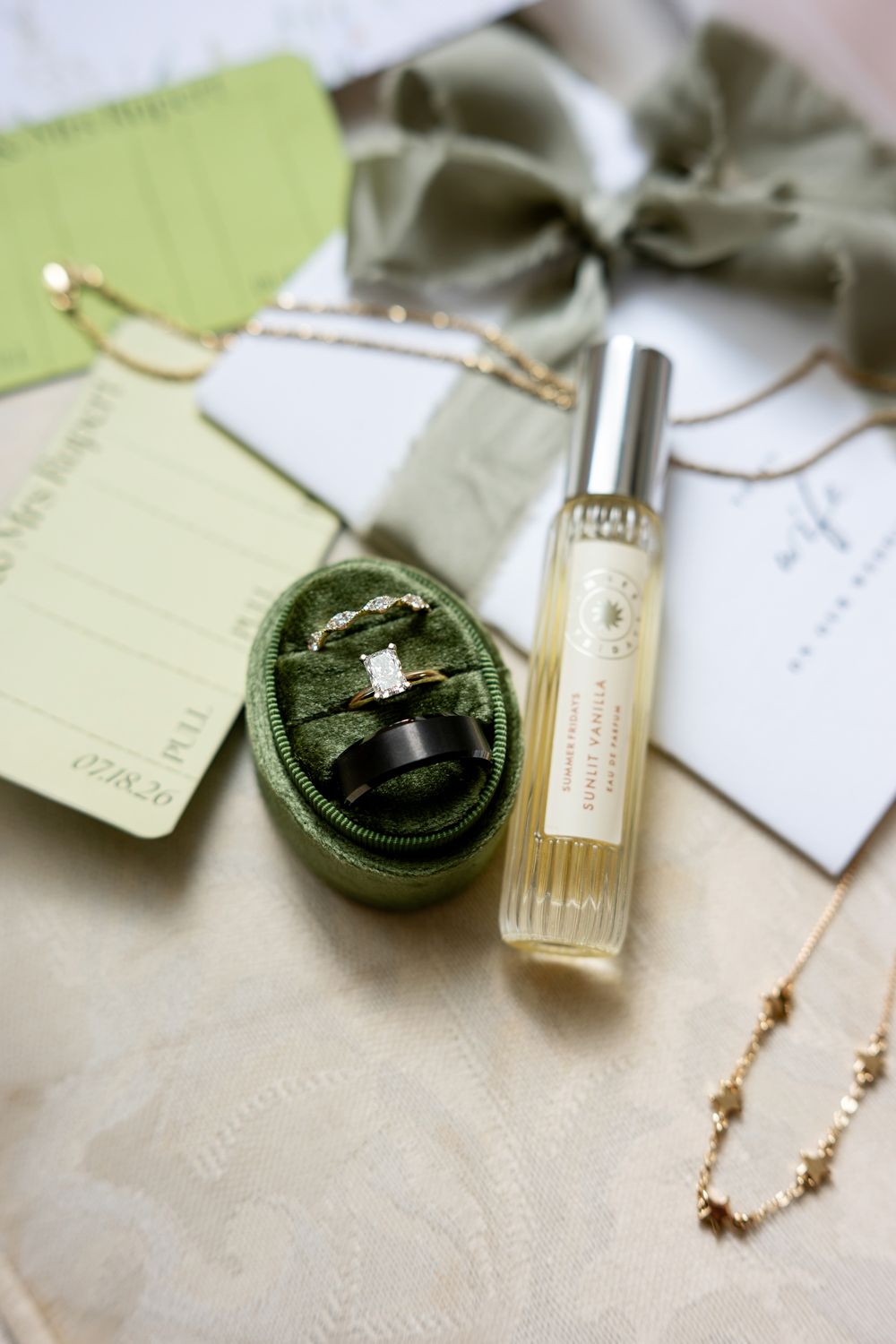 Green velvet ring box with wedding bands, perfume roller, gold necklace, and sage ribbon on elegant cream surface.