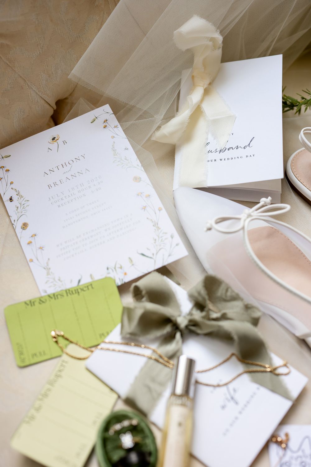 Elegant wedding flat lay with floral invitation, sage green ribbon, perfume, jewelry, and bridal heels on neutral background.