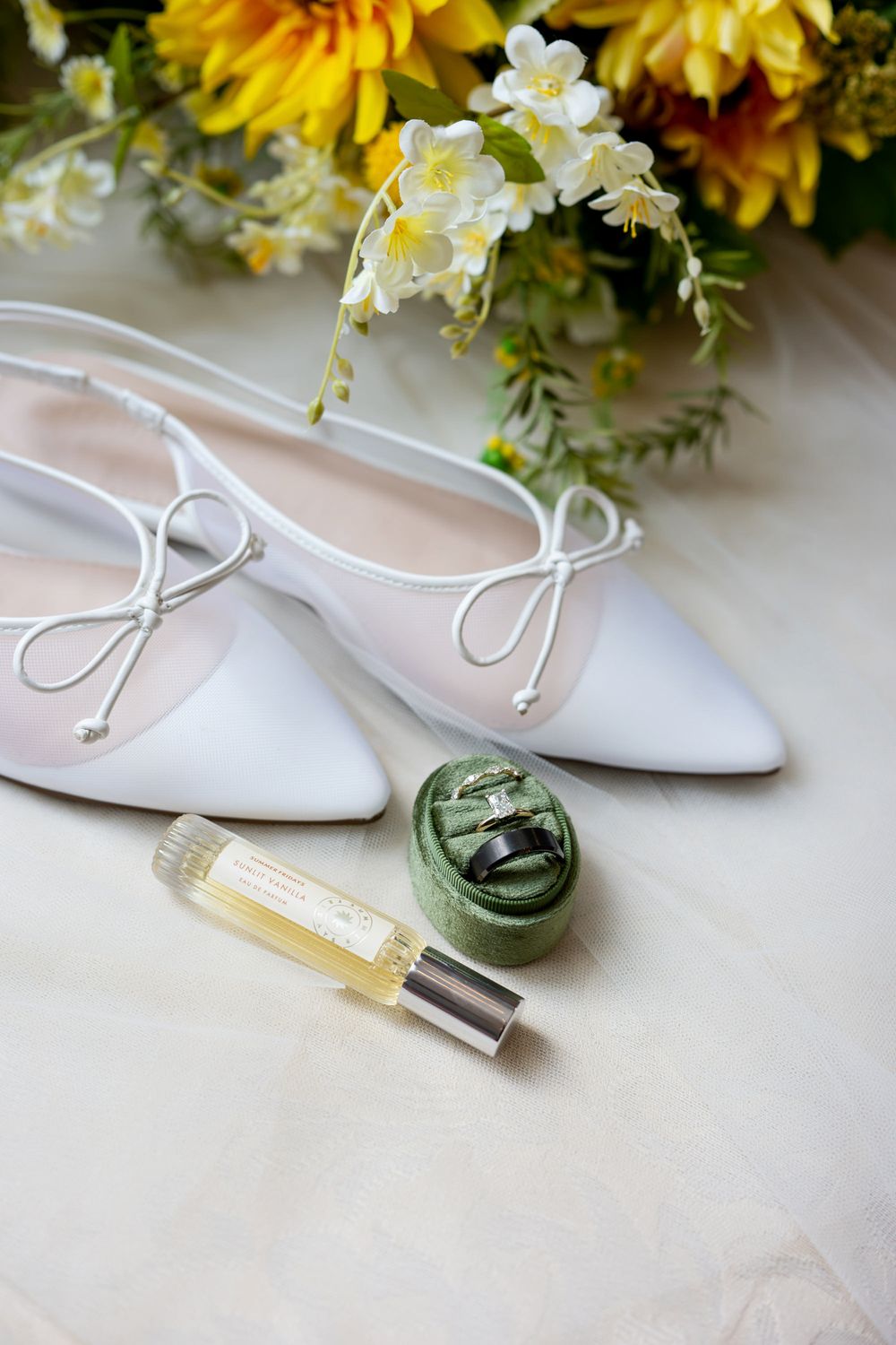 White bridal flats with bow ties beside green velvet ring box, perfume roller, and yellow sunflower bouquet on ivory surface.