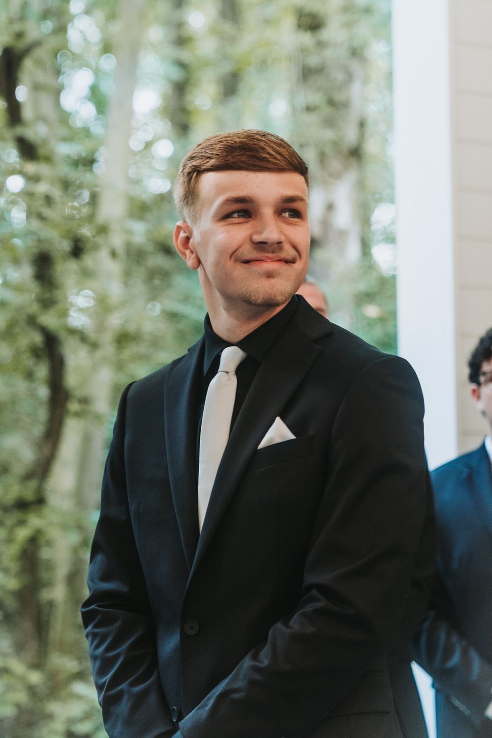 Young man in black tuxedo with white tie and pocket square smiling at a wedding ceremony outdoors.