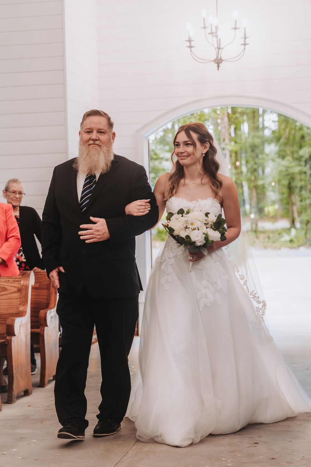 Bride in white ballgown walks down chapel aisle with bearded man in black suit, holding white floral bouquet.