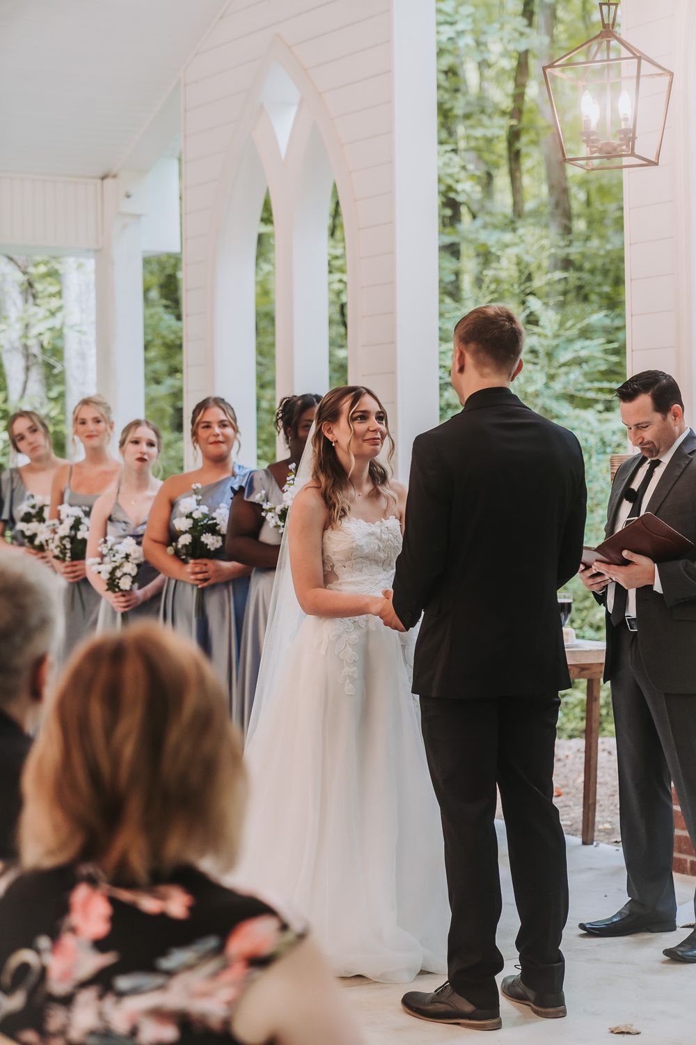 Bride and groom exchange vows at outdoor wedding ceremony with bridesmaids in blue gowns under white arched pavilion.