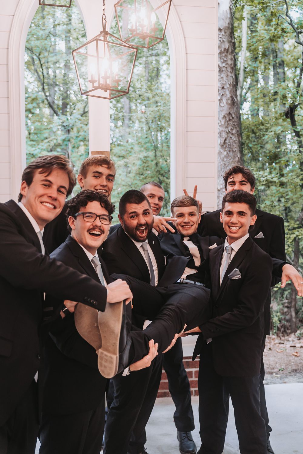 Groomsmen in black suits lifting one man while posing and laughing outdoors near white columns and lanterns.