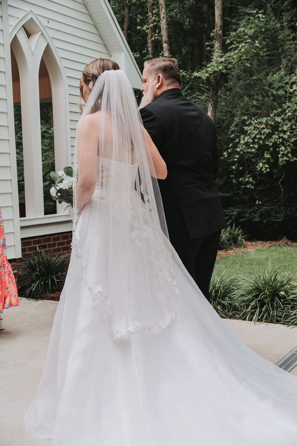 Bride in white gown and veil stands with groom in black suit outside a white chapel surrounded by trees.