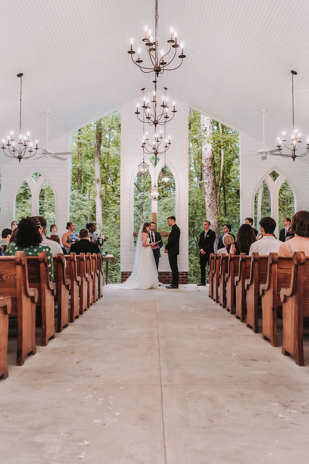 Elegant open-air chapel wedding ceremony with wooden pews, chandelier lighting, and couple exchanging vows at the altar.