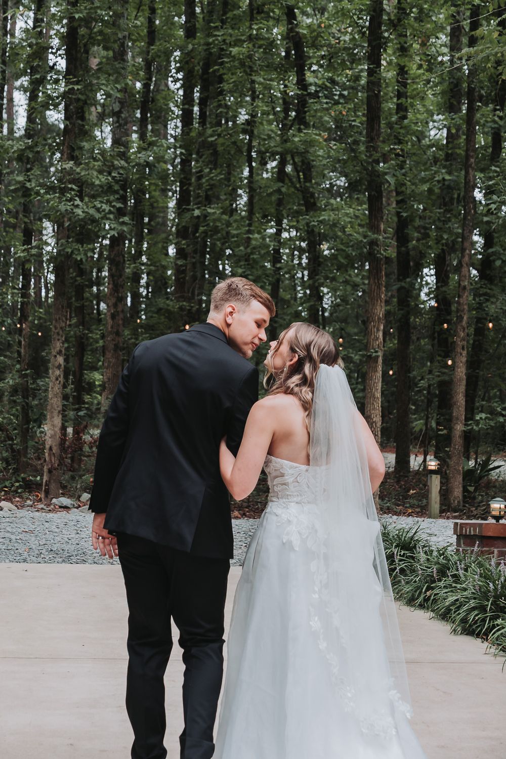 A bride and groom share an intimate moment, holding hands on a paved path surrounded by tall trees at their outdoor wedding.