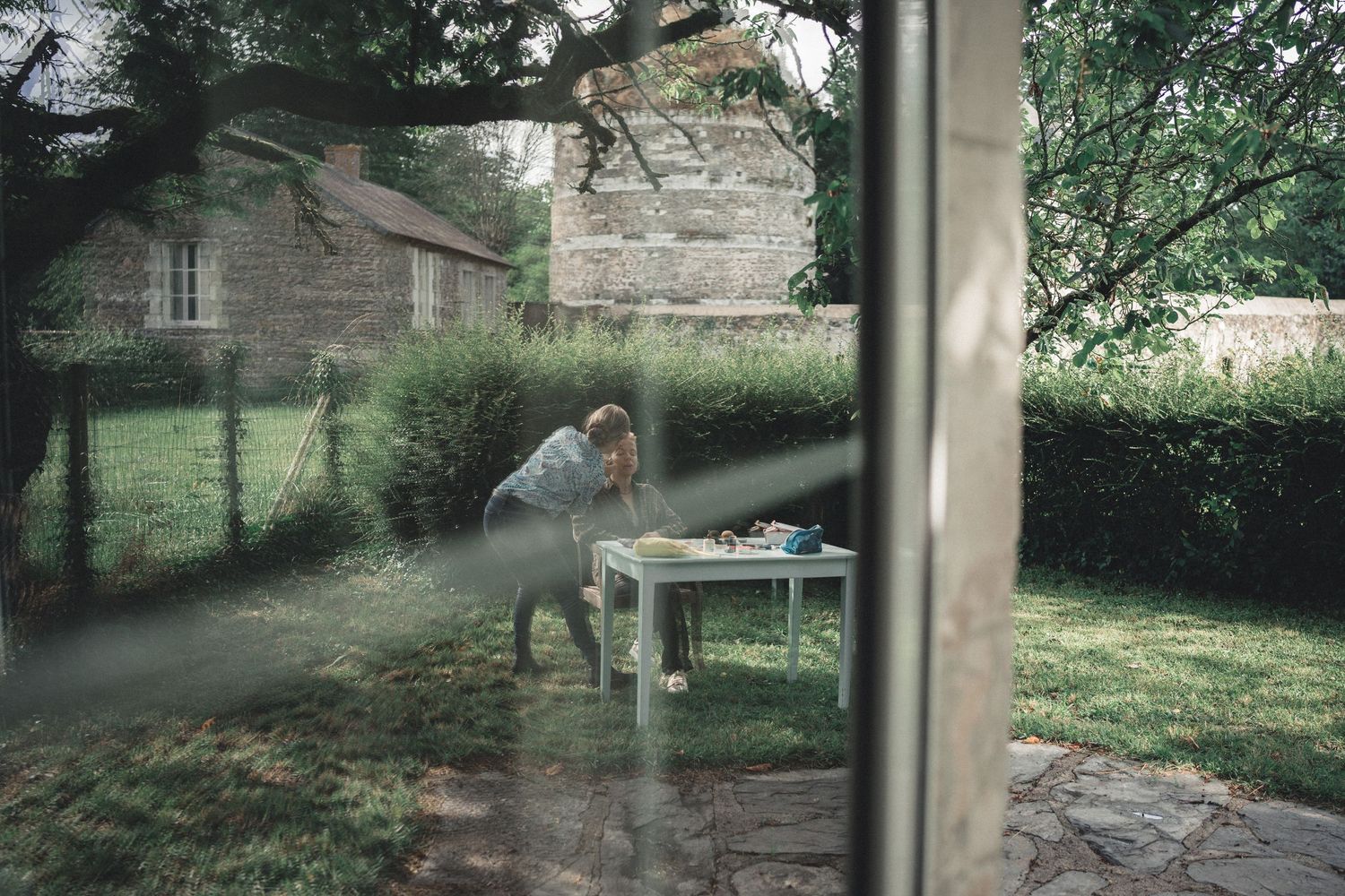 Person sitting at a white table in a lush garden, viewed through a window.