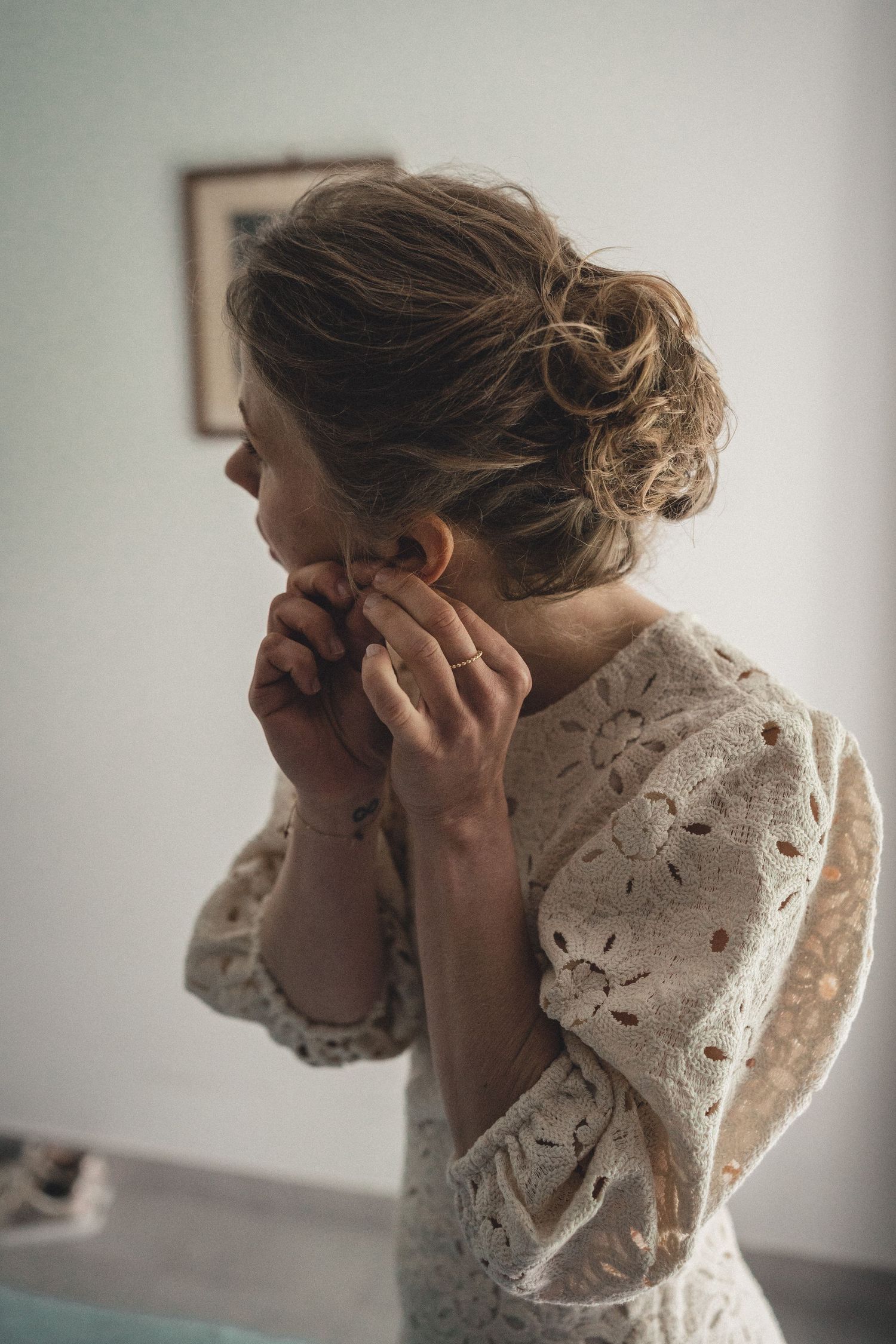 Bride in lace dress putting on earring before wedding ceremony.
