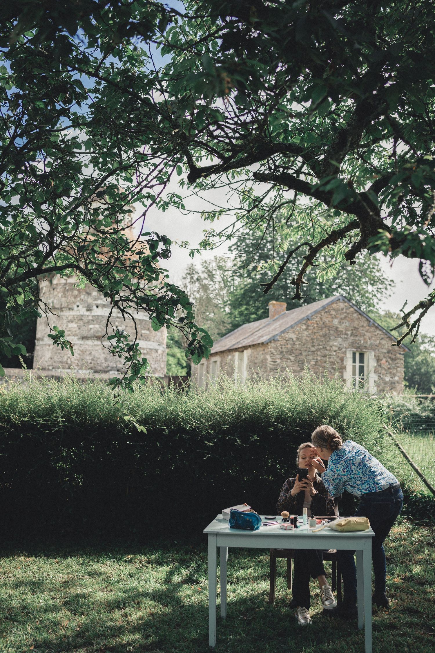 Two people share an intimate moment at a white outdoor table beneath a large tree at a rustic stone property.