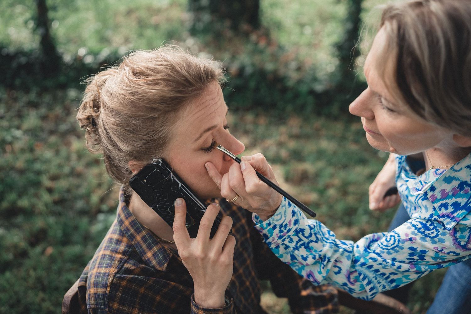 Woman talking on phone while another applies eye makeup outdoors in a natural wooded setting.