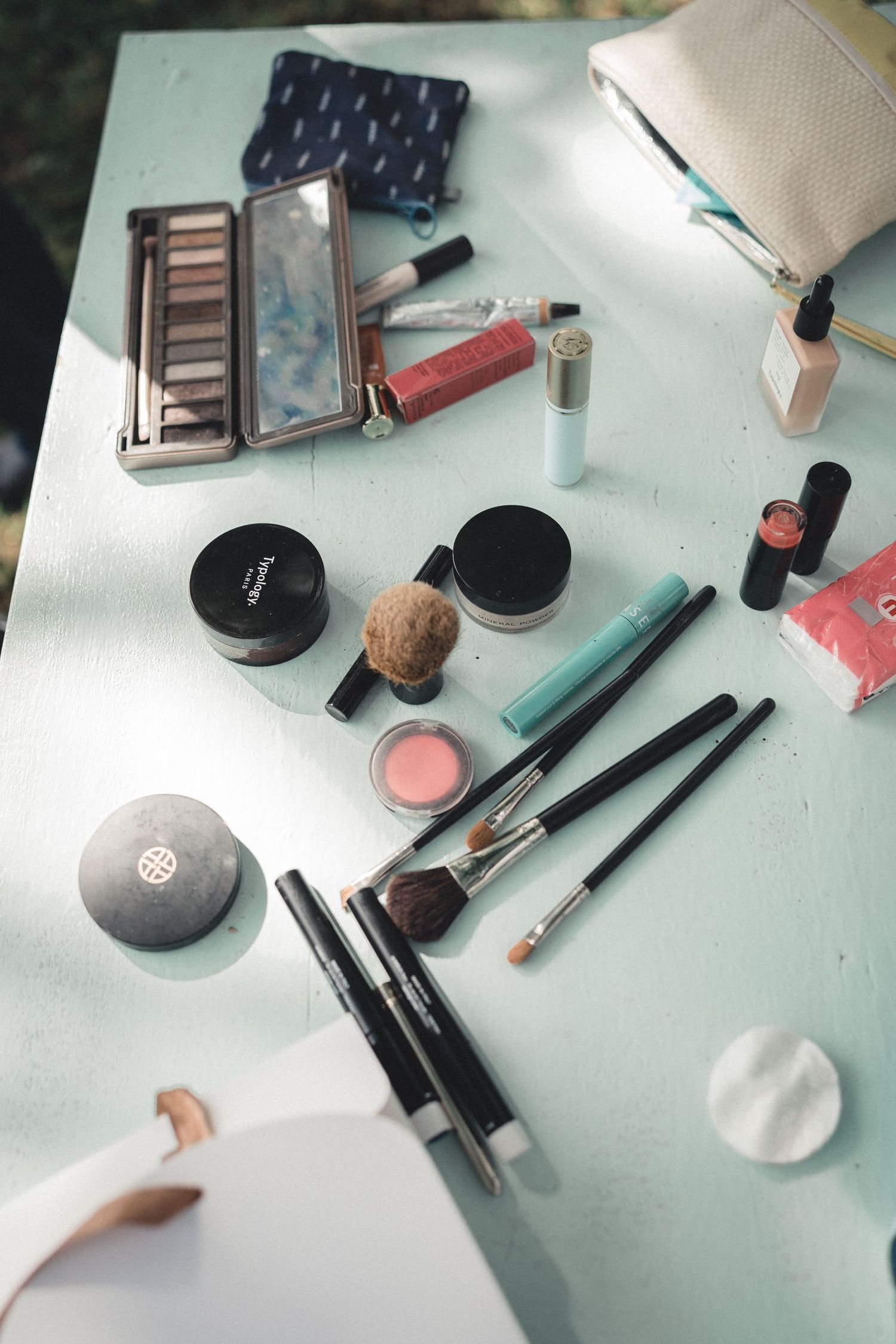 Wedding makeup flatlay with brushes, eyeshadow palette, lipstick, and cosmetics on light blue table.