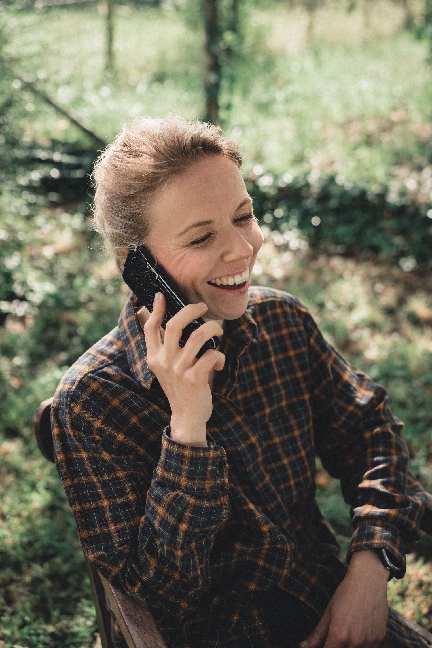 A cheerful woman in a plaid shirt laughs while talking on her smartphone in a lush green setting.