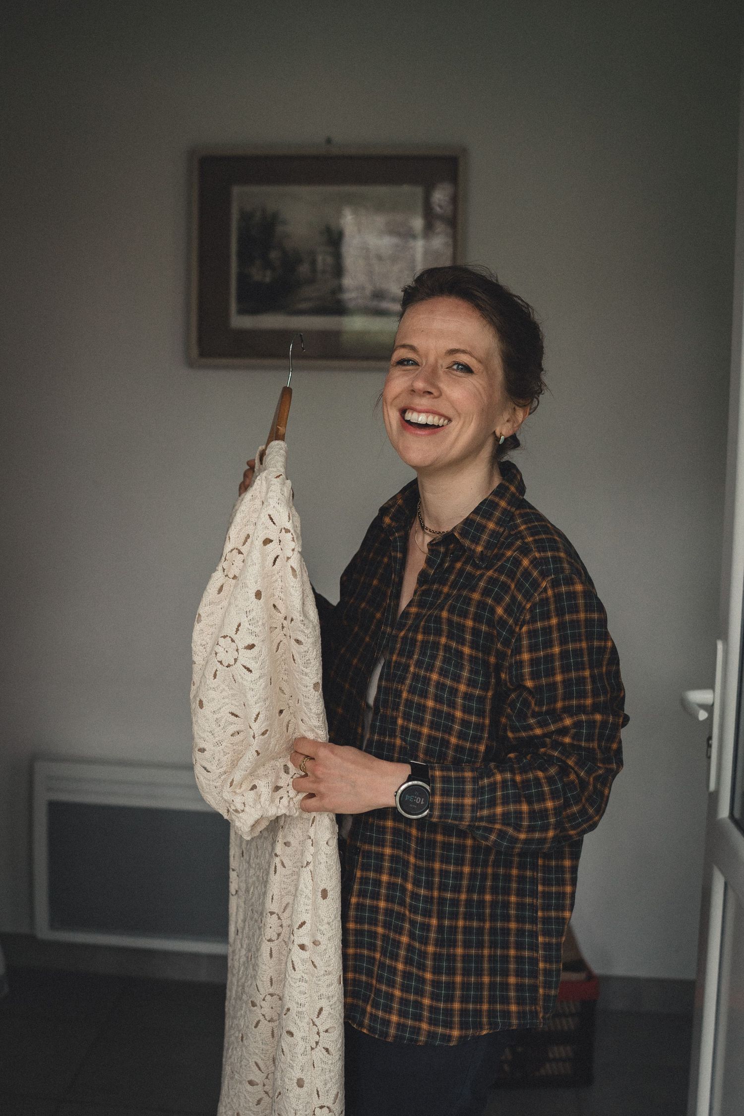 Smiling bride holding a white lace wedding dress on a hanger in a cozy room.