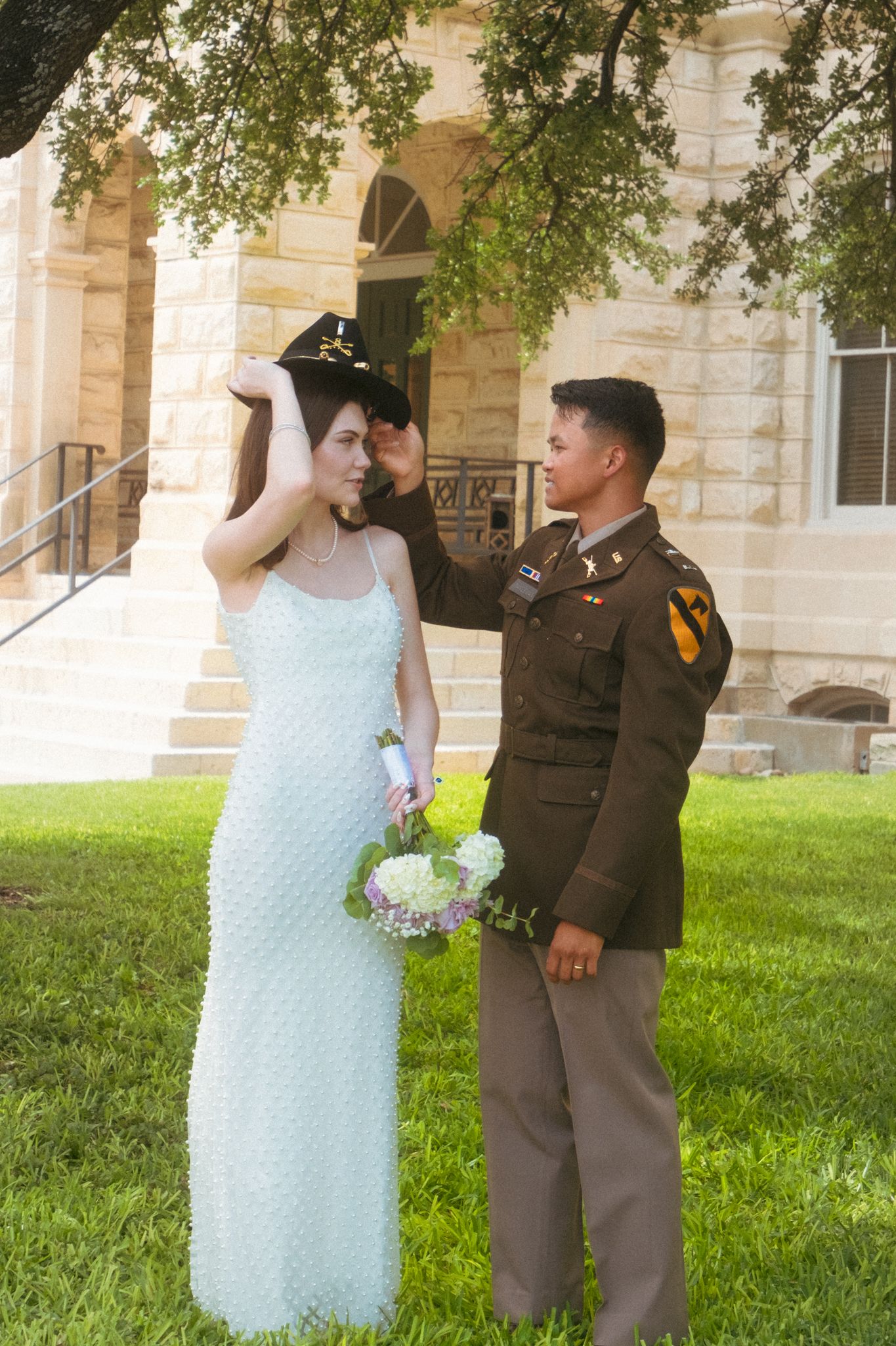 Military groom placing hat on bride in white dress outside stone building Belton Courthouse for wedding photography in Belton Texas