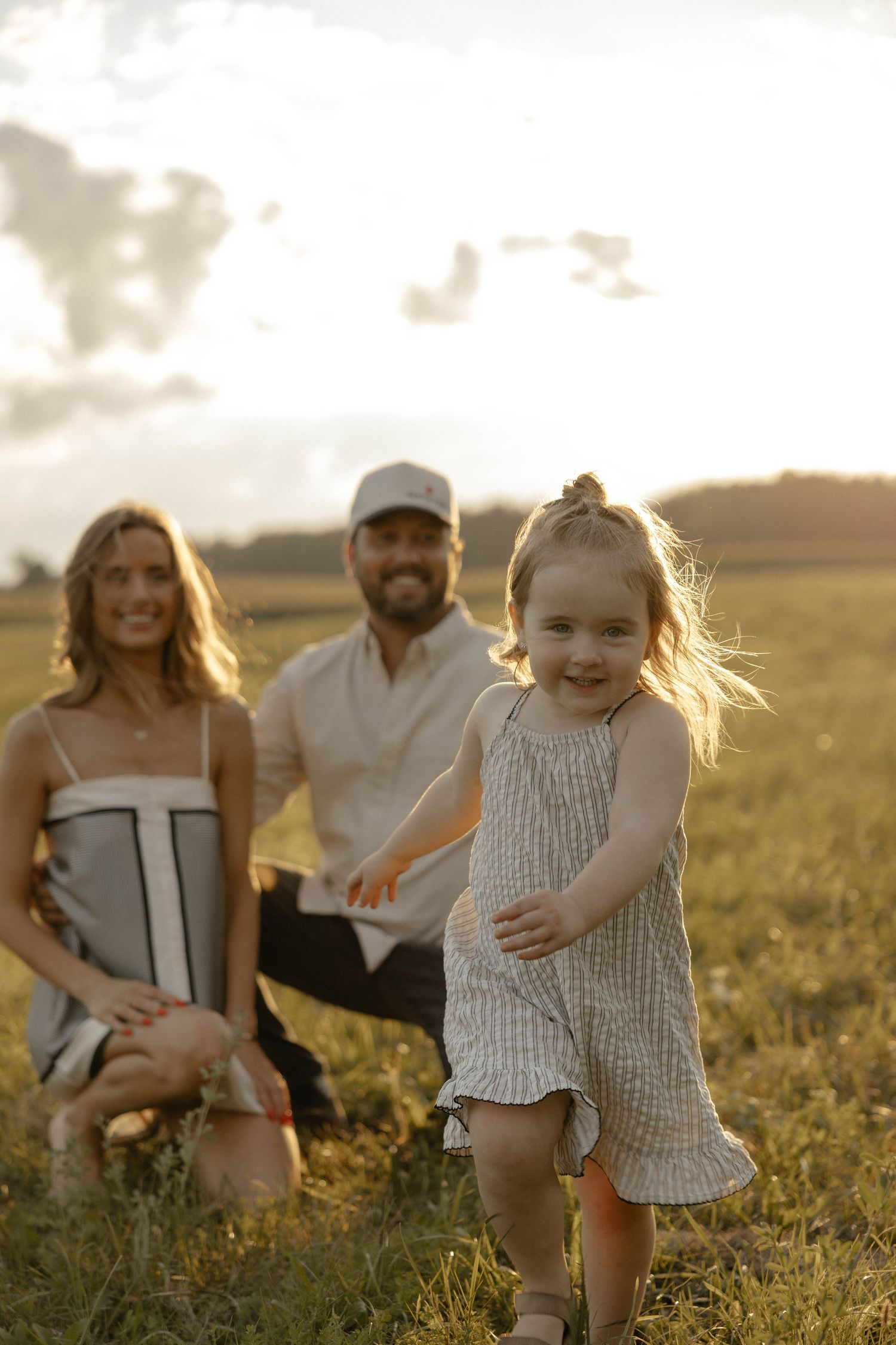 A young girl in a striped dress runs through a golden field at sunset while her smiling parents kneel in the background.