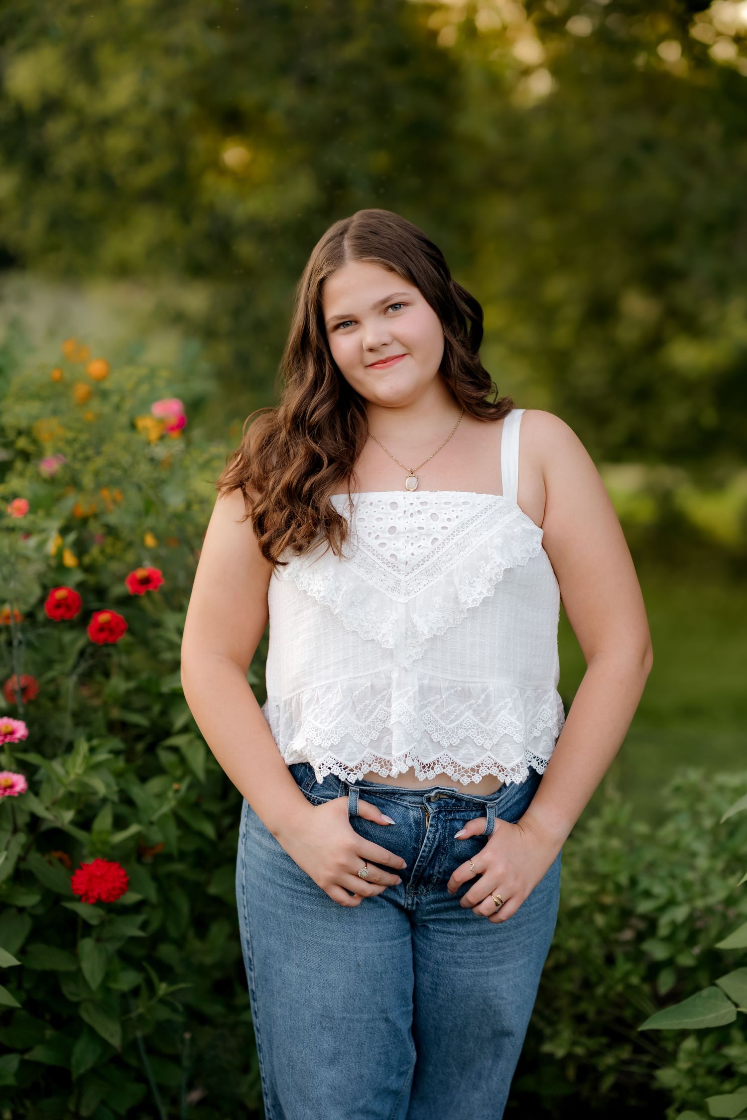 Teen girl in white lace crop top and jeans posing outdoors among colorful garden flowers.