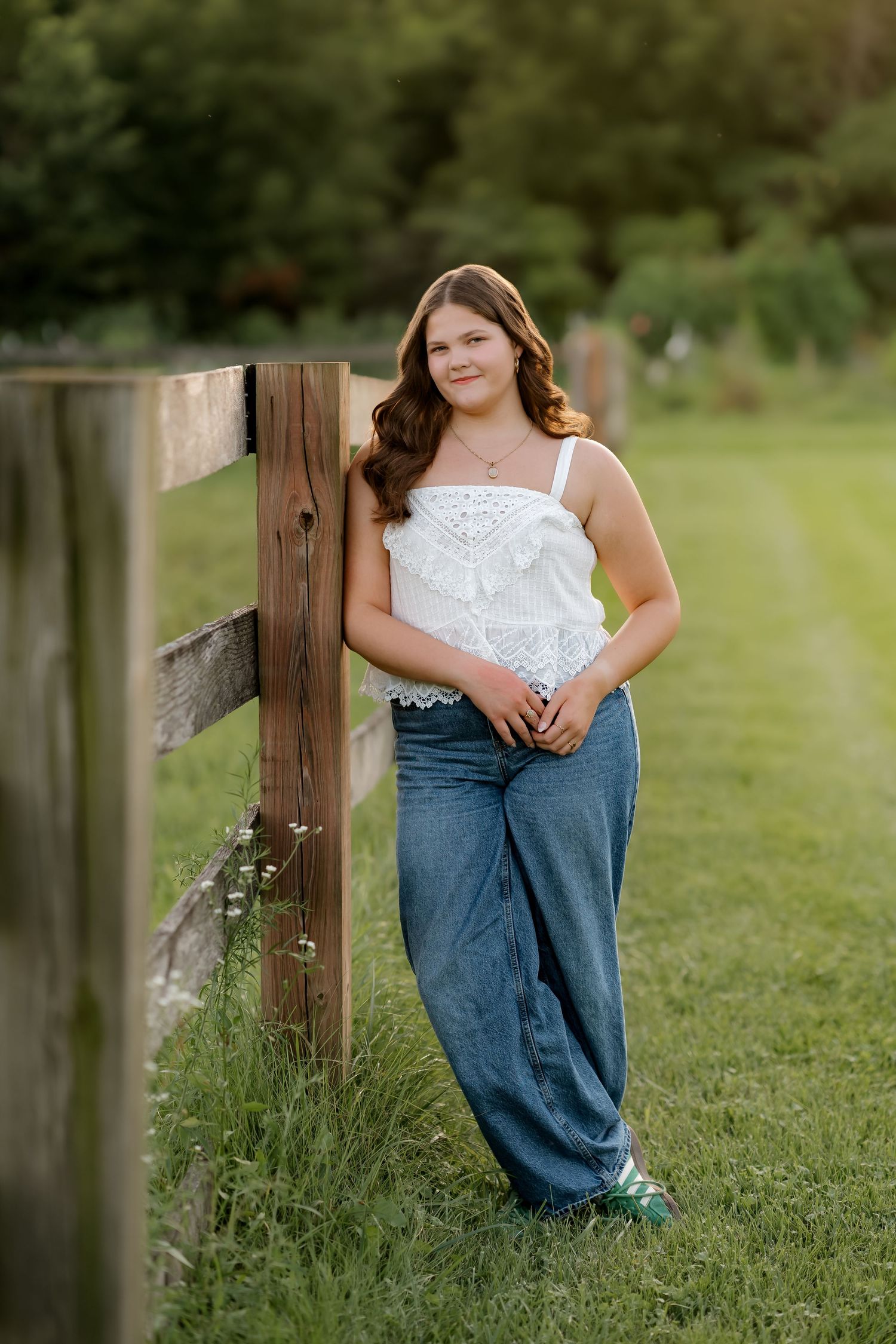 Young woman in white lace top and wide-leg jeans leaning against a wooden fence post in a green field.