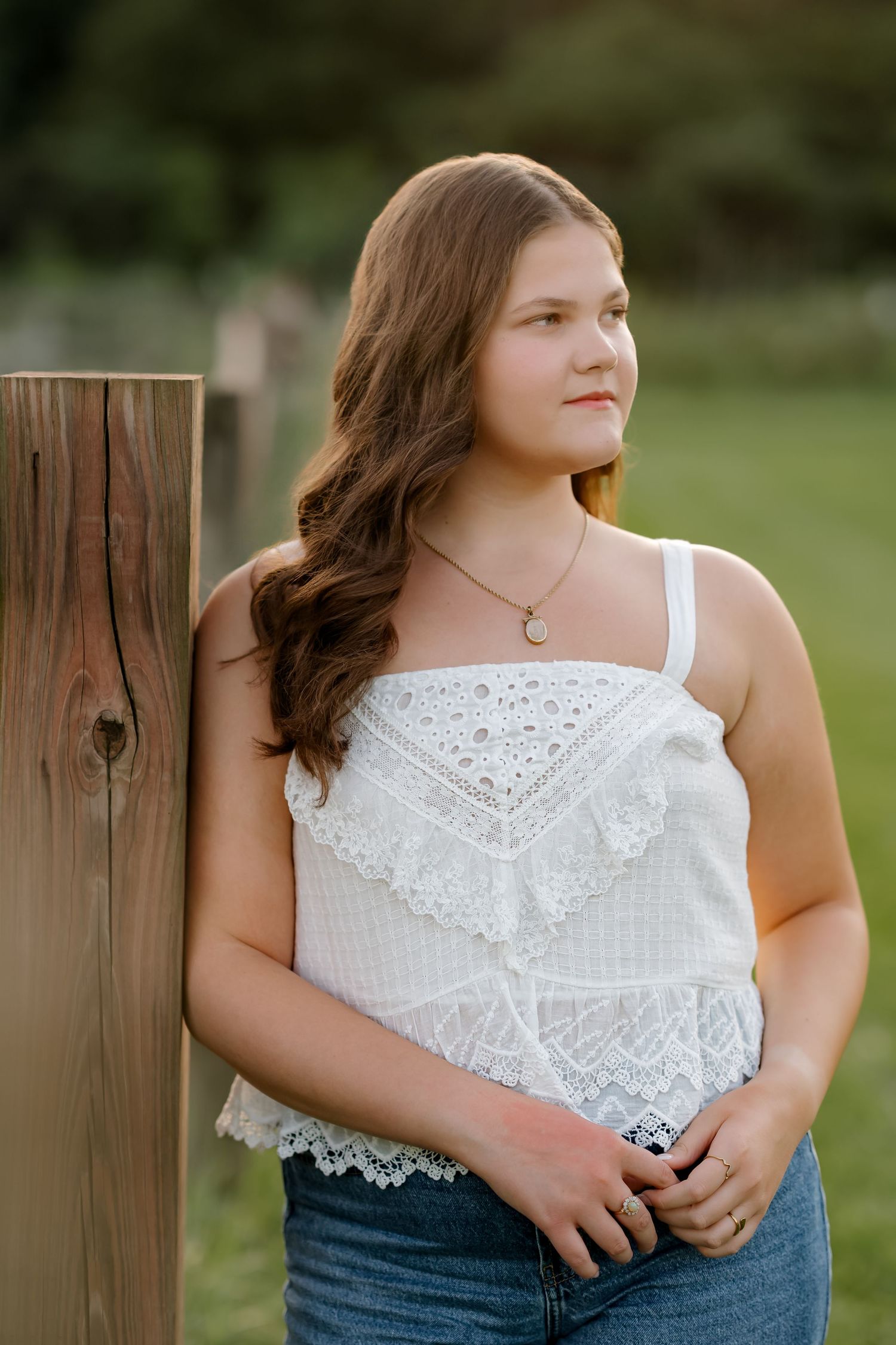 Young woman in white lace top and jeans leaning against a wooden post outdoors in a portrait photo.