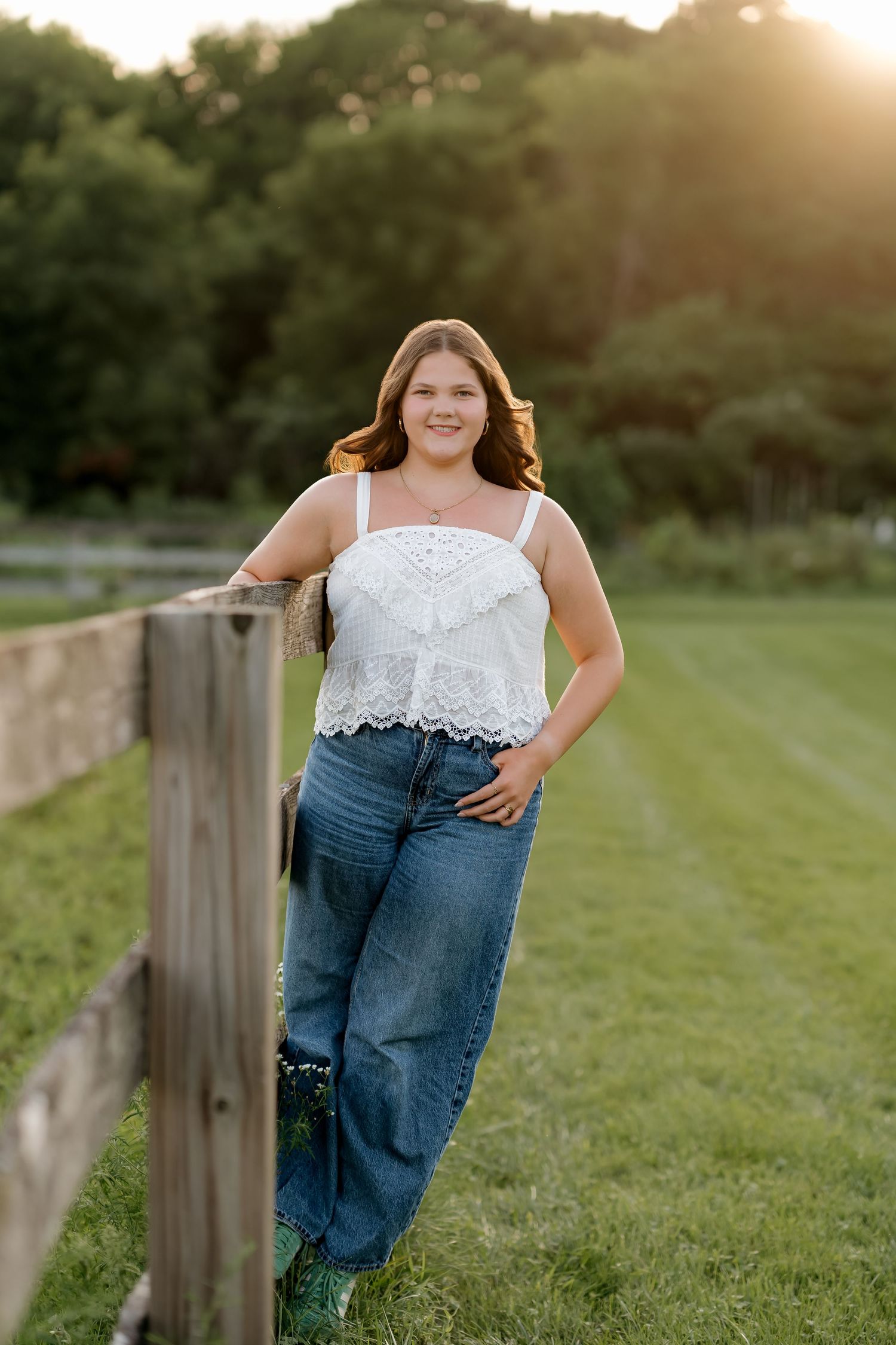 Young woman in white lace top and jeans leaning on wooden fence at golden hour in green countryside.