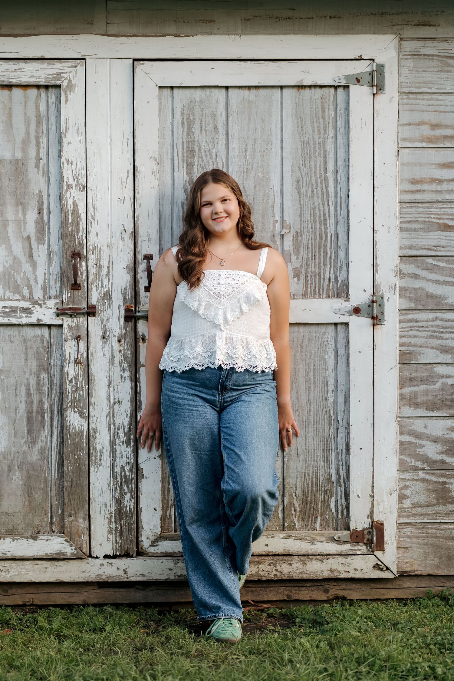 Young woman in white lace tank top and jeans smiling against rustic weathered barn doors outdoors.