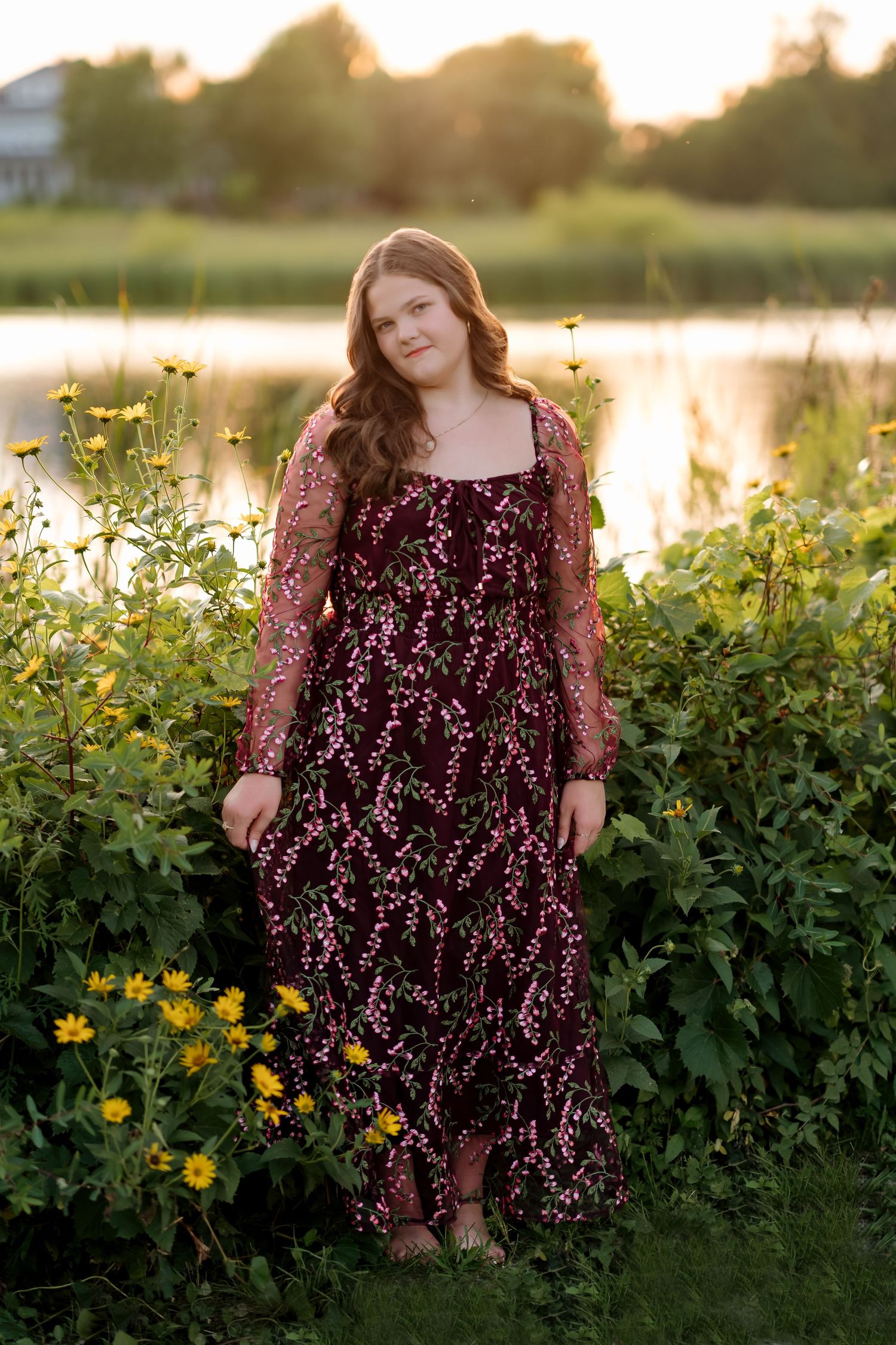 Young woman in floral burgundy dress standing among wildflowers near a pond at golden hour.