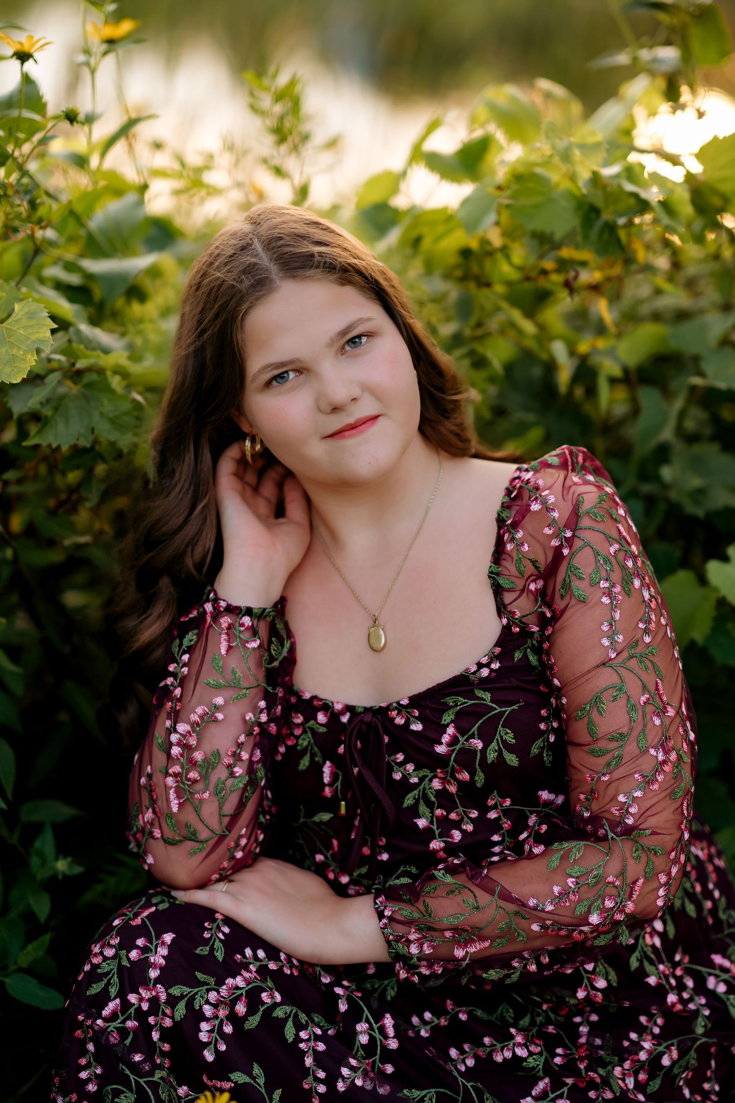Young woman in floral burgundy dress poses gracefully among green foliage in golden hour light.