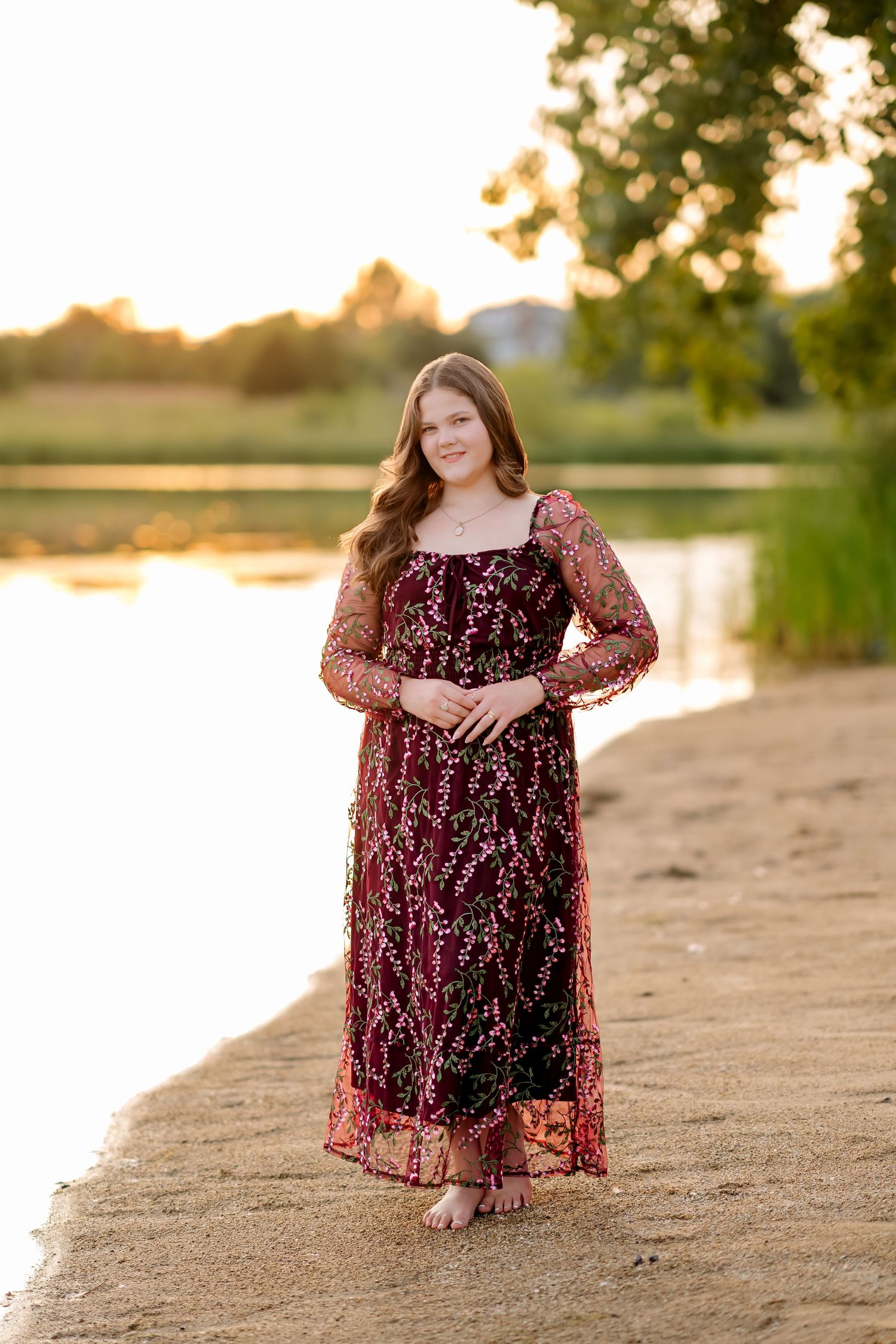 Young woman in burgundy floral lace gown standing barefoot by a sunlit lake at golden hour.