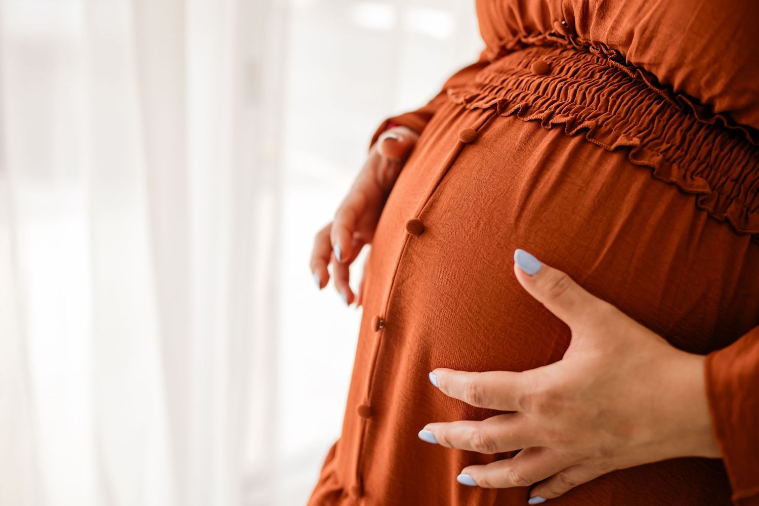 Pregnant woman in rust orange dress cradling her baby bump with both hands near a bright window.