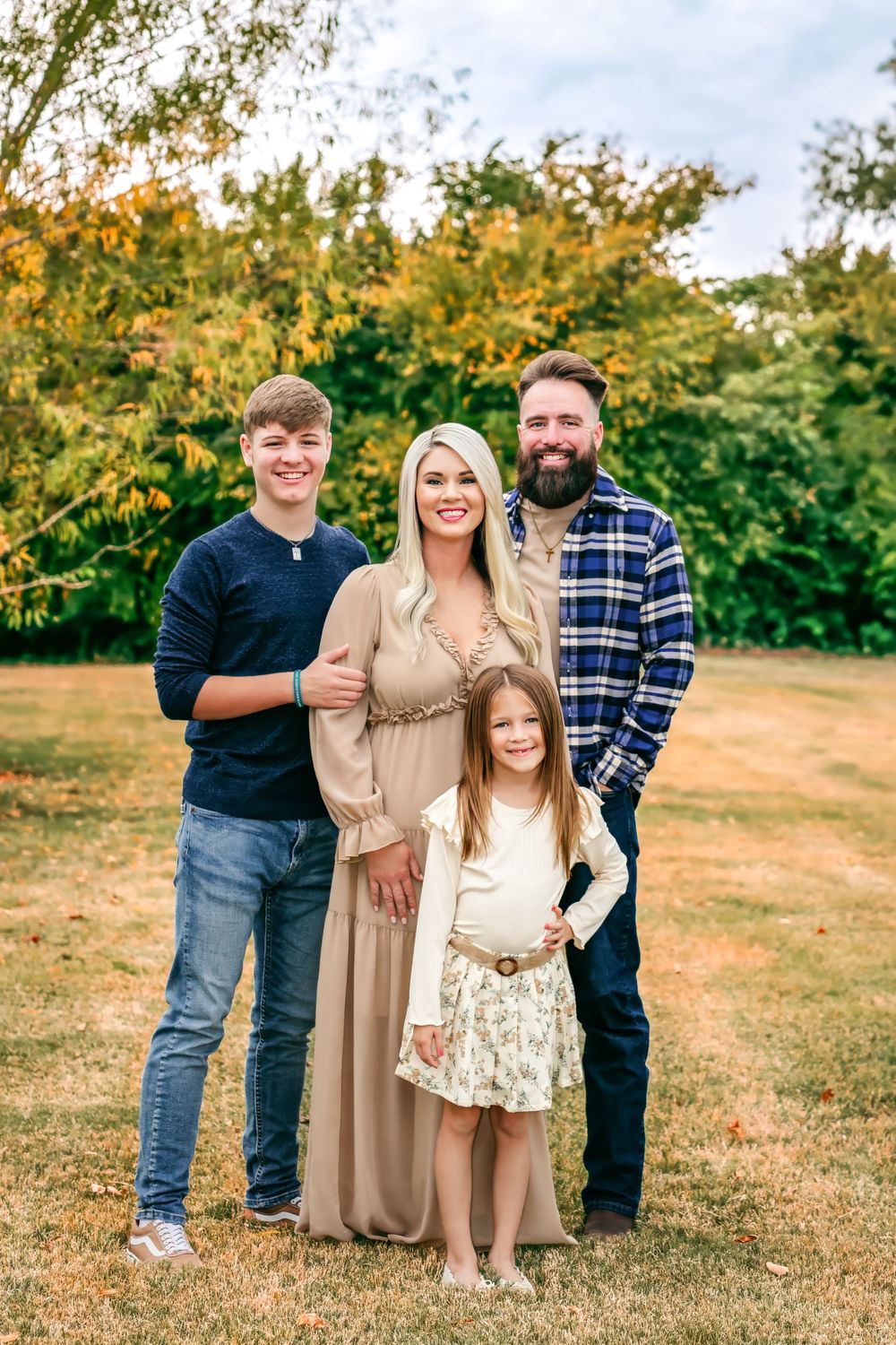 Family of four posing outdoors in autumn park, pregnant mother in tan dress with bearded father and two children.