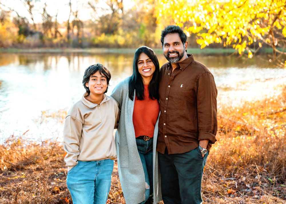 Family of three smiling outdoors near a river with golden autumn foliage in the background.