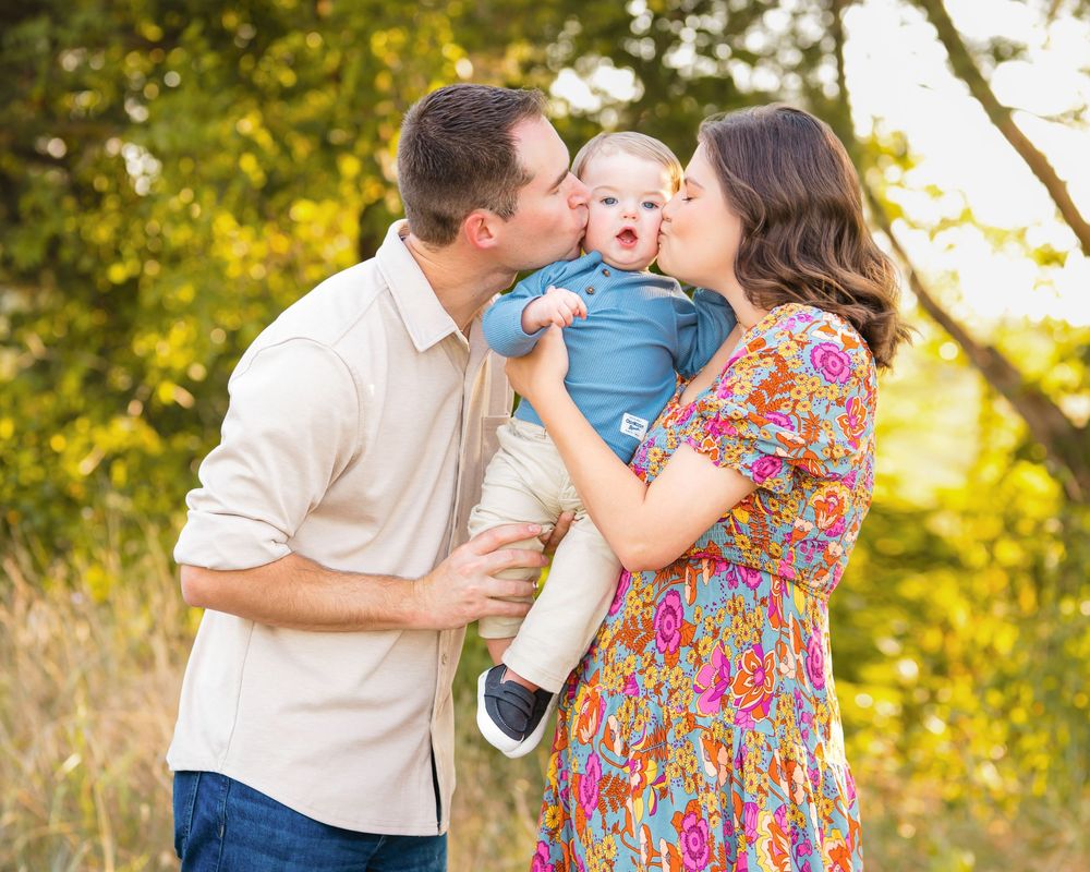 A happy couple kisses their smiling baby outdoors among golden trees in a warm family portrait.