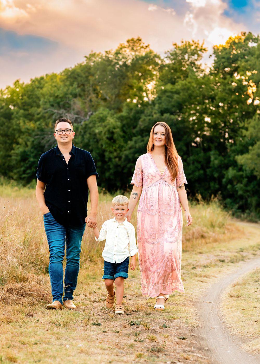 Pregnant woman in pink dress walks with partner and young son on a scenic outdoor path at golden hour.