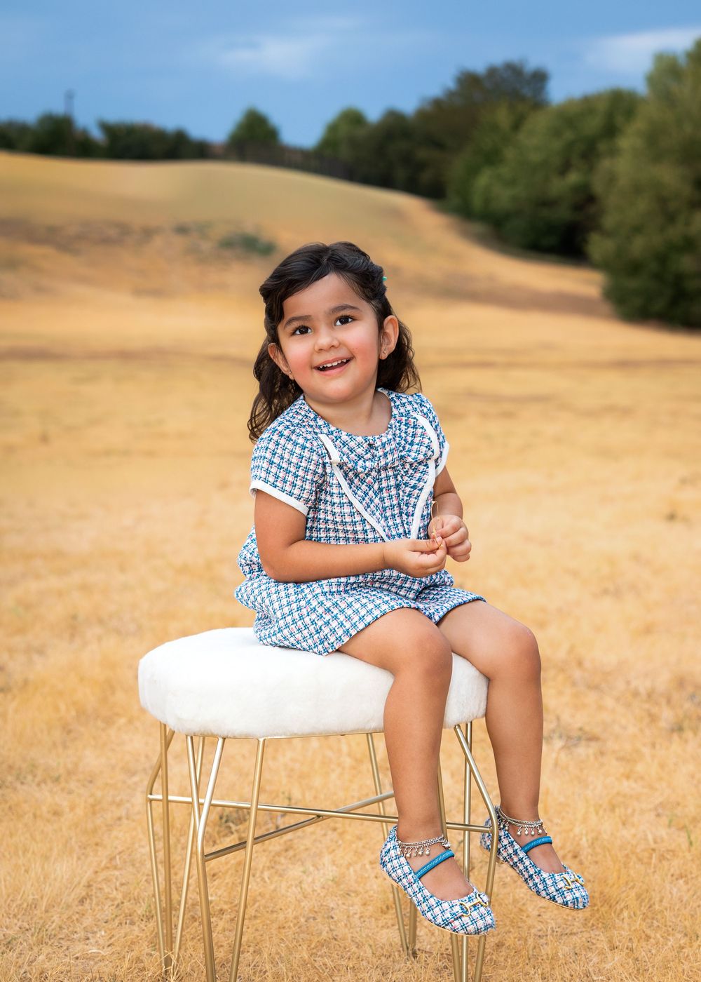 Young girl in blue gingham outfit sits on white stool in golden field, smiling happily outdoors.