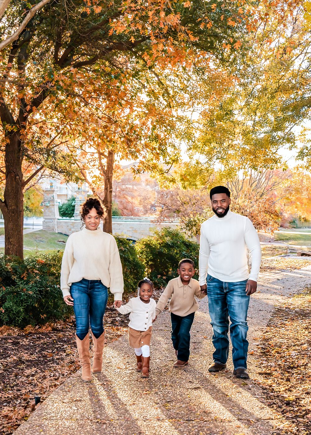 Family of four walking outdoors in fall foliage, parents holding children's hands on a tree-lined path.