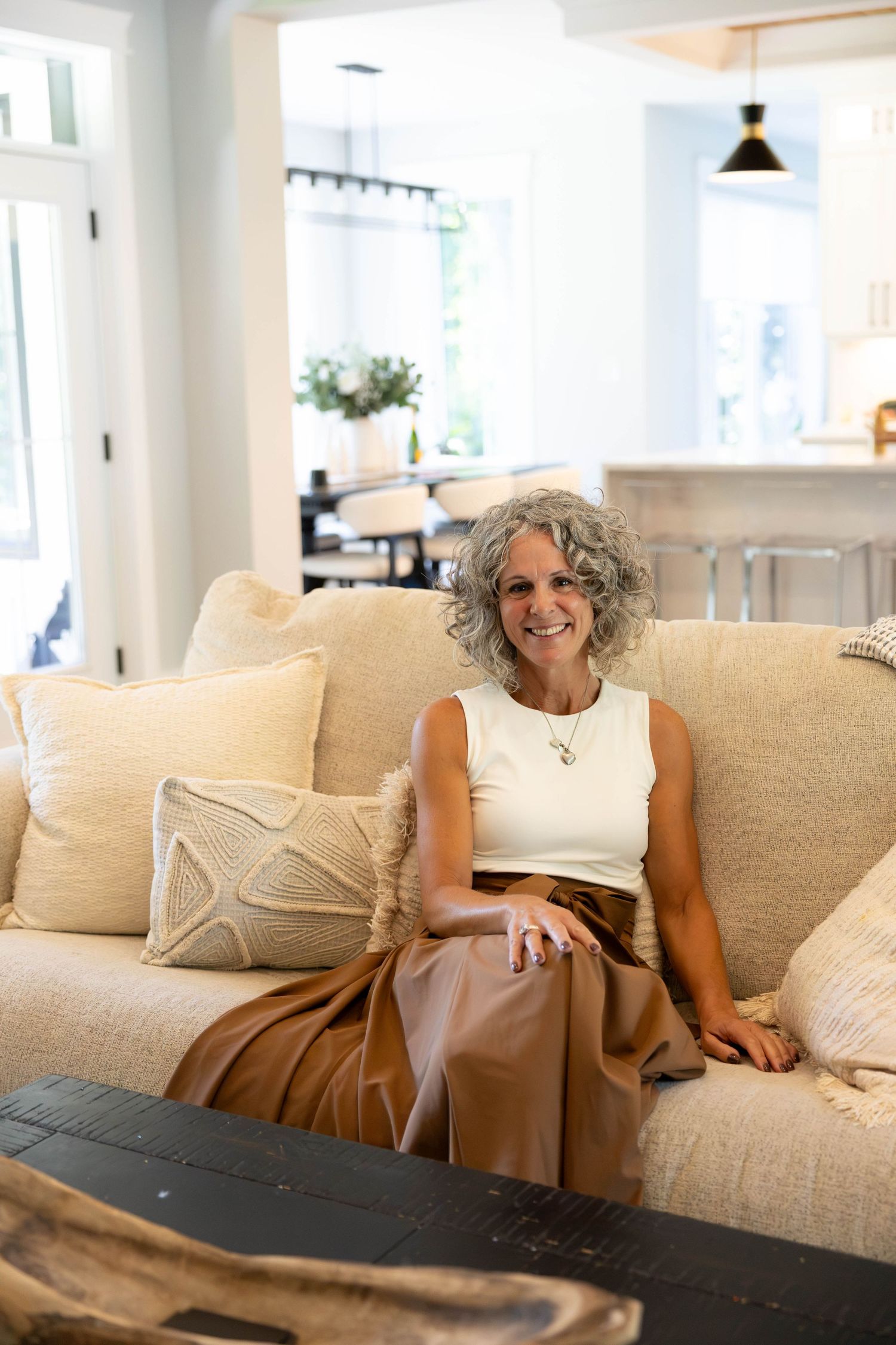 Smiling woman with gray curly hair sits on a beige sofa in a bright, modern living room wearing a white top.