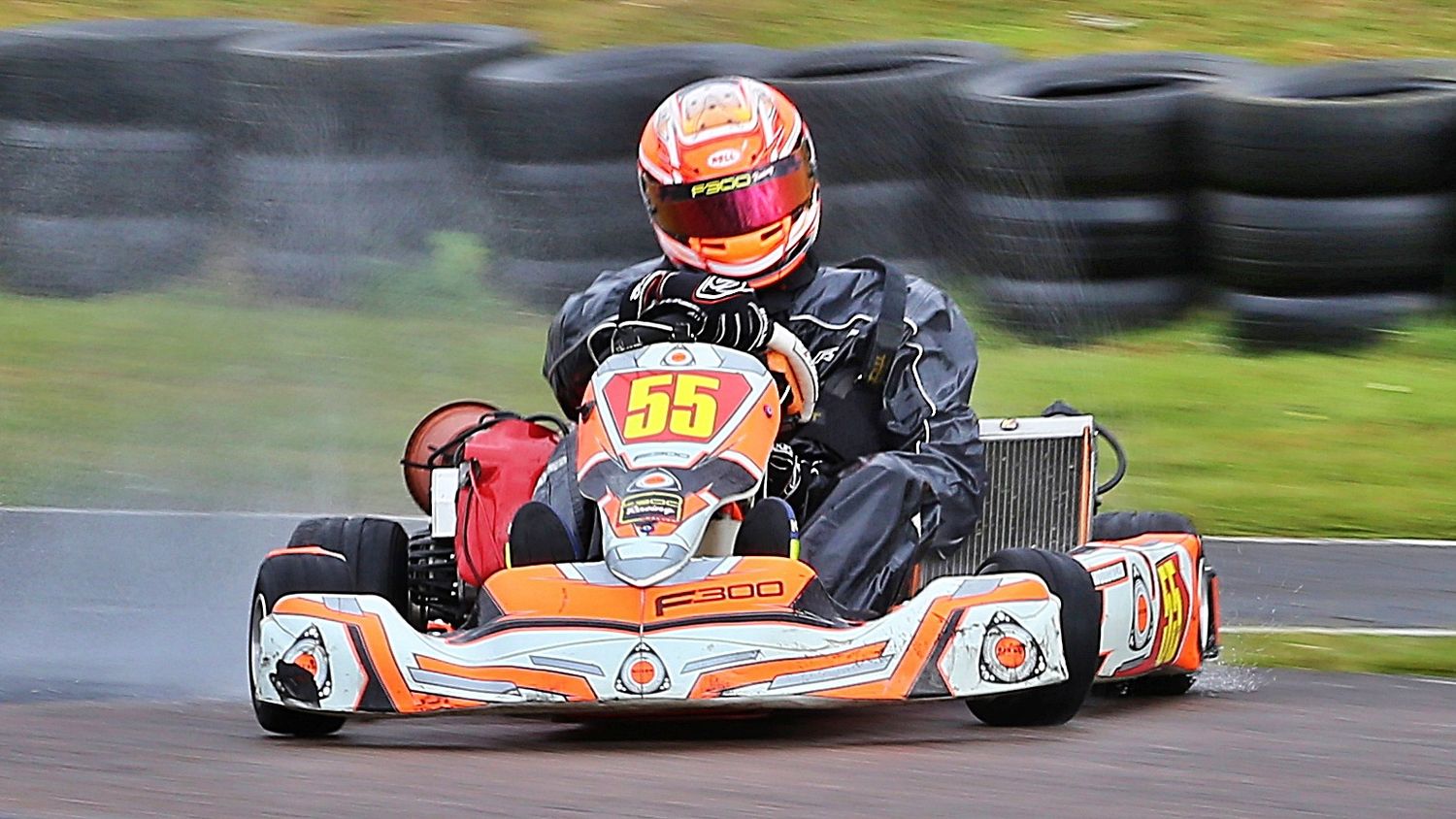 Go-kart number 55 in orange and white racing on a wet track in rainy conditions.