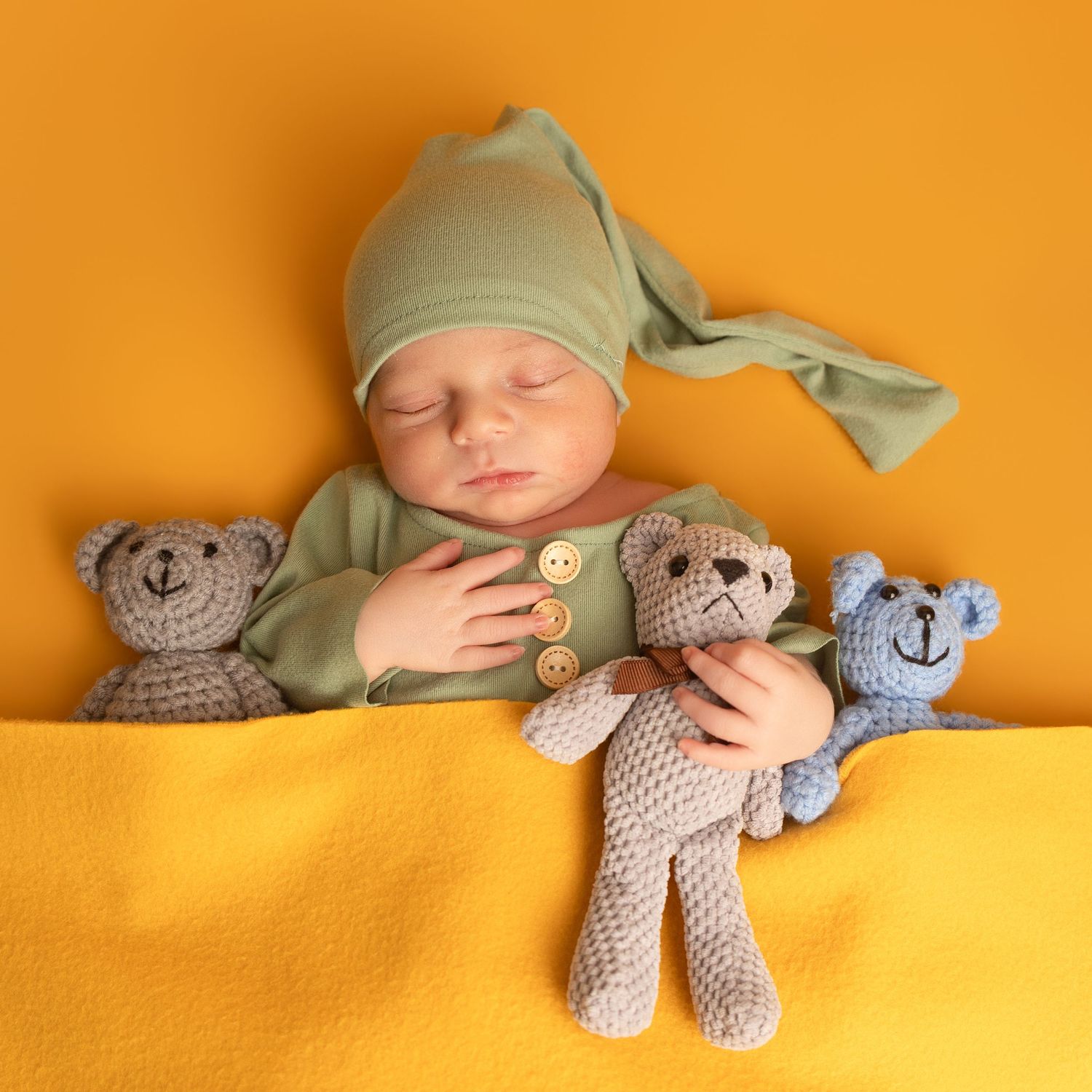 Sleeping newborn baby in green outfit and knotted hat cuddles three crocheted teddy bears on yellow blanket.