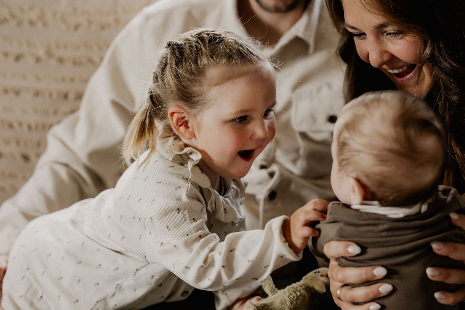 Ein kleines Mädchen lächelt ein Baby an, das von einer lachenden Frau gehalten wird, daneben ein Mann in einem warmen Familienmoment.