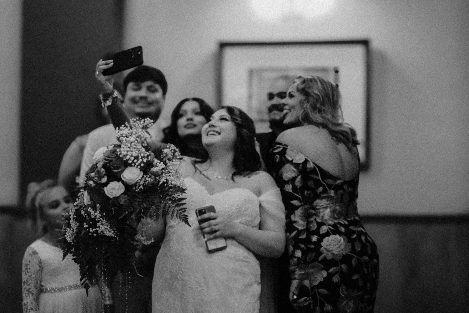 Black and white photo of a smiling bride in a white gown holding bouquet and drink while taking a group selfie at wedding reception.