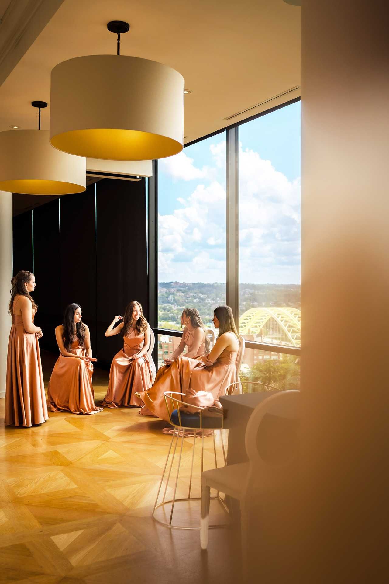 Bridesmaids in blush pink gowns gather by floor-to-ceiling windows with scenic views, at the view wedding venue