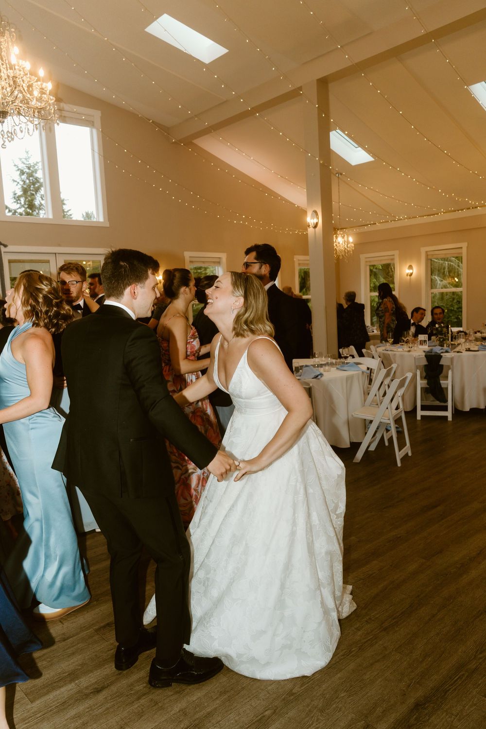 Bride and groom share a joyful first dance at elegant wedding reception with fairy lights and chandeliers.
