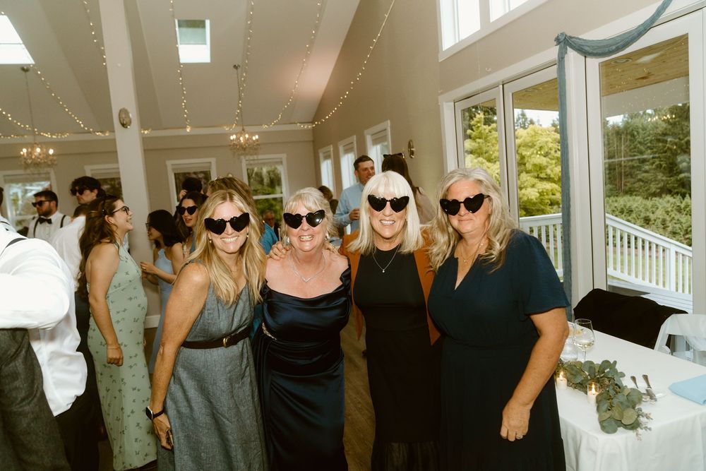 Four women wearing sunglasses smile together at an elegant indoor wedding reception venue.