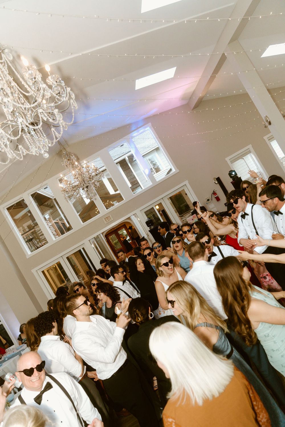 Crowded wedding reception dance floor with guests celebrating under chandeliers and string lights in elegant white venue.