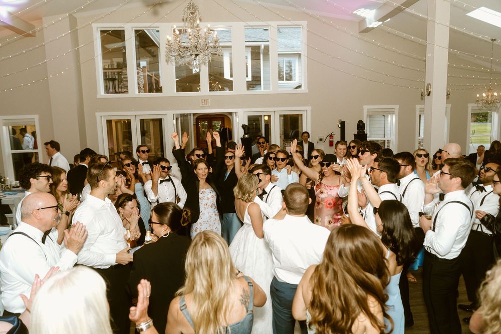 Guests celebrating at a wedding reception, dancing in an elegant white venue with chandelier lighting.